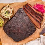 A beef brisket on a board with some slices cut off, next to pickled onions, coleslaw and pickles