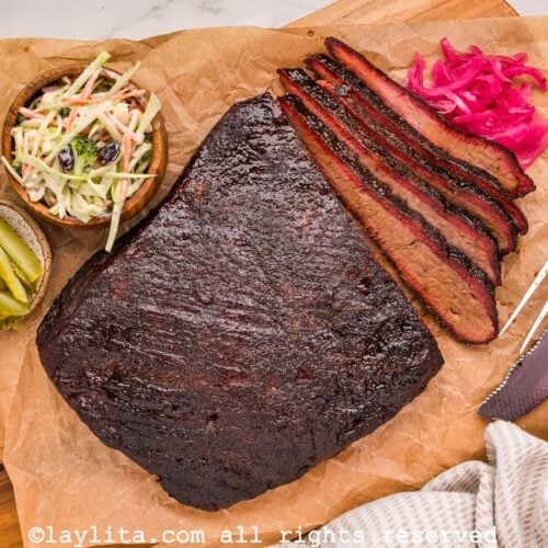 A beef brisket on a board with some slices cut off, next to pickled onions, coleslaw and pickles