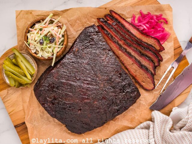 A beef brisket on a board with some slices cut off, next to pickled onions, coleslaw and pickles