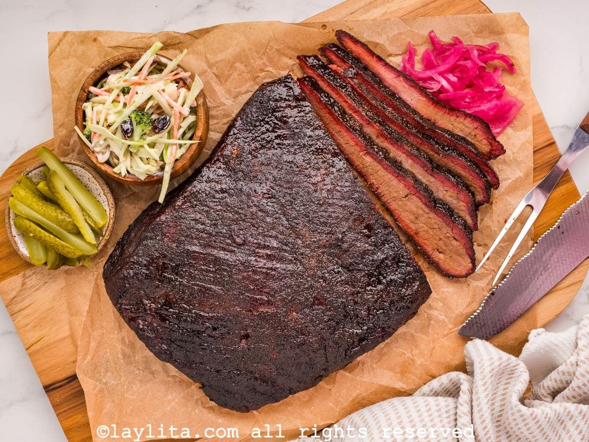 A beef brisket on a board with some slices cut off, next to pickled onions, coleslaw and pickles