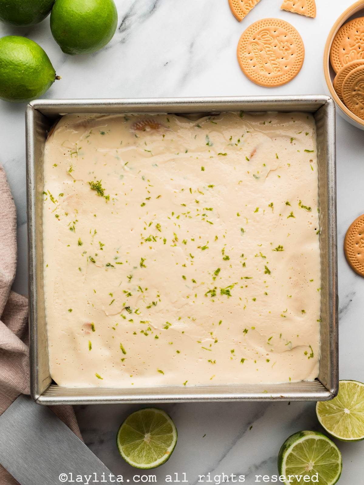 Overhead view of a square pan filled with carlota de limon dessert, with lime zest on top, there are limes and cookies scattered on the sides
