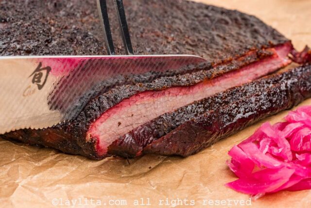 Knife slicing into finished smoked brisket next to pickled onions