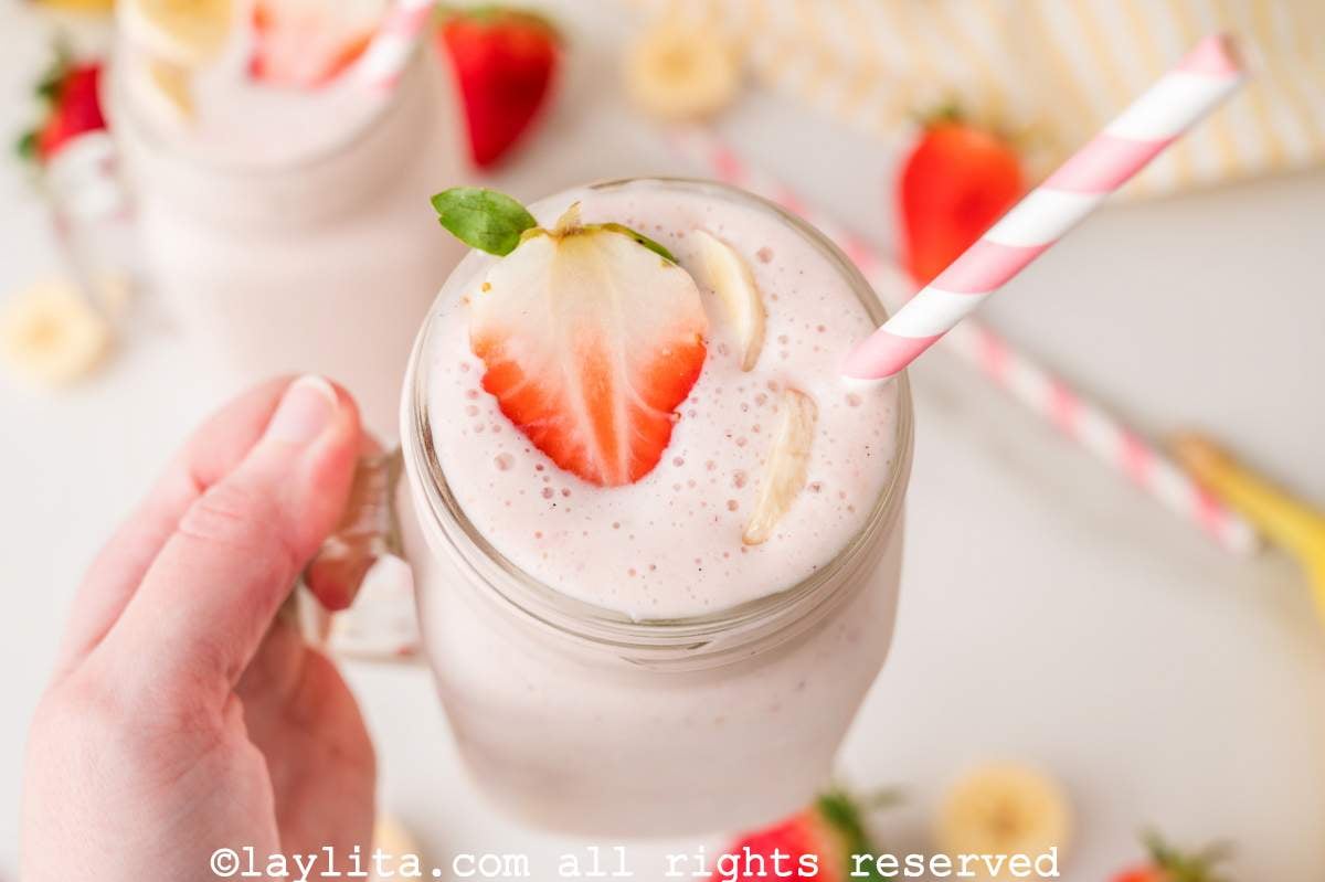 Hand holding a strawberry banana milkshake that is garnished with whipped cream and fresh fruit