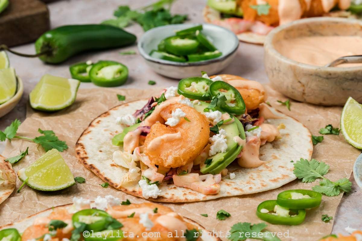 Side view of an open baja shrimp taco on brown parchment paper, with a bowl of sauce and sliced jalapeños in the background