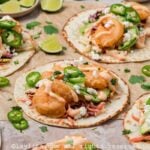 Side view of several Baja shrimp tacos on brown parchment paper topped with sauce and sliced jalapeños with lime slices on the side