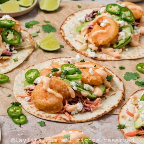 Side view of several Baja shrimp tacos on brown parchment paper topped with sauce and sliced jalapeños with lime slices on the side