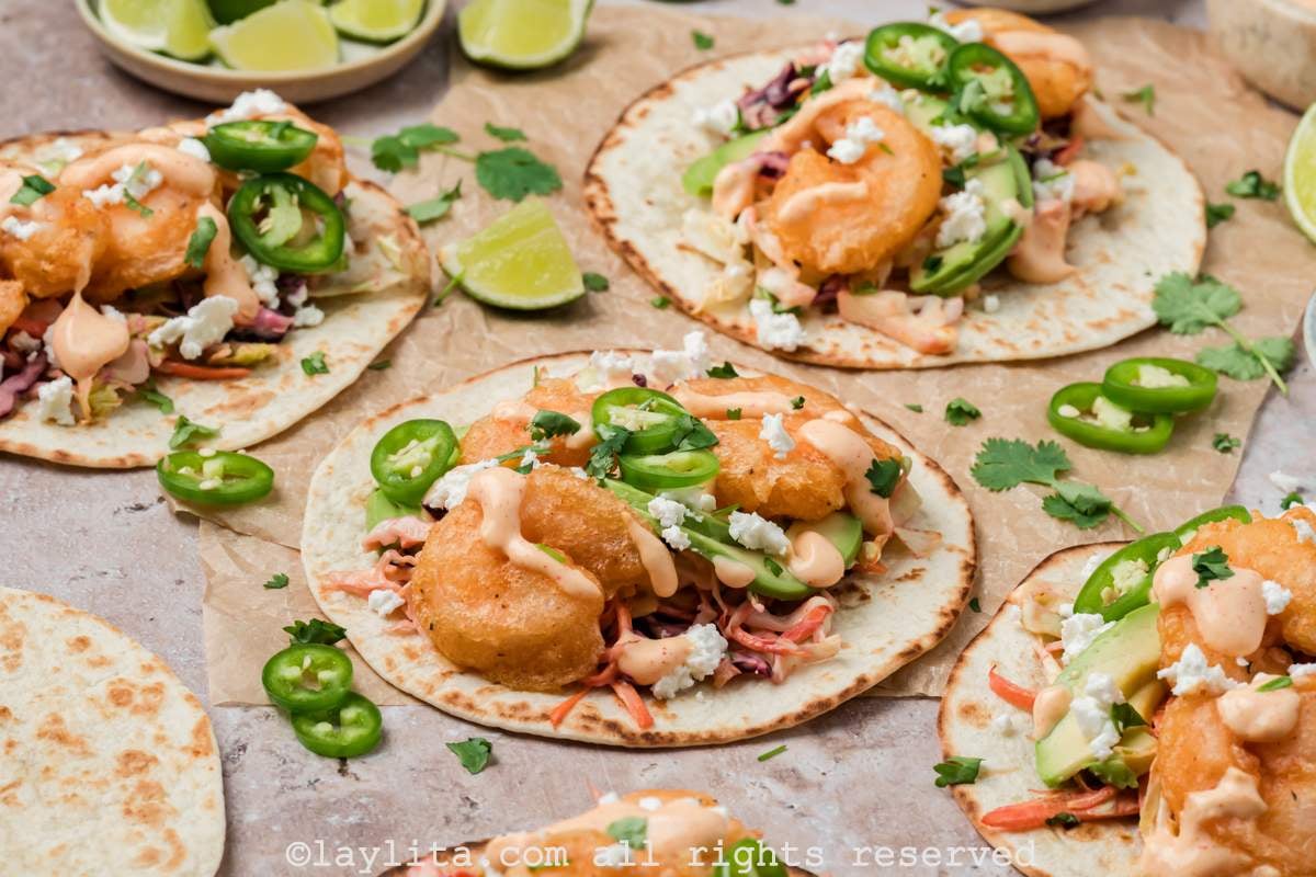 Side view of several Baja shrimp tacos on brown parchment paper topped with sauce and sliced jalapeños with lime slices on the side