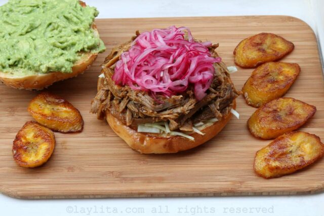 A pulled pork sandwich topped with pickled onions and the top part of the sandwich on the side and fried plantains on the side