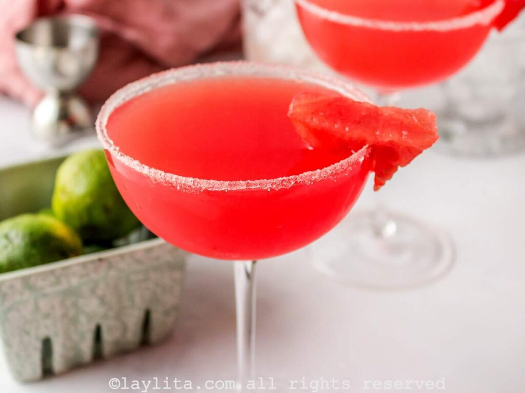 Side view of two watermelon margaritas in coupe glasses, both garnished with sugar and watermelon, with a container of limes in the background