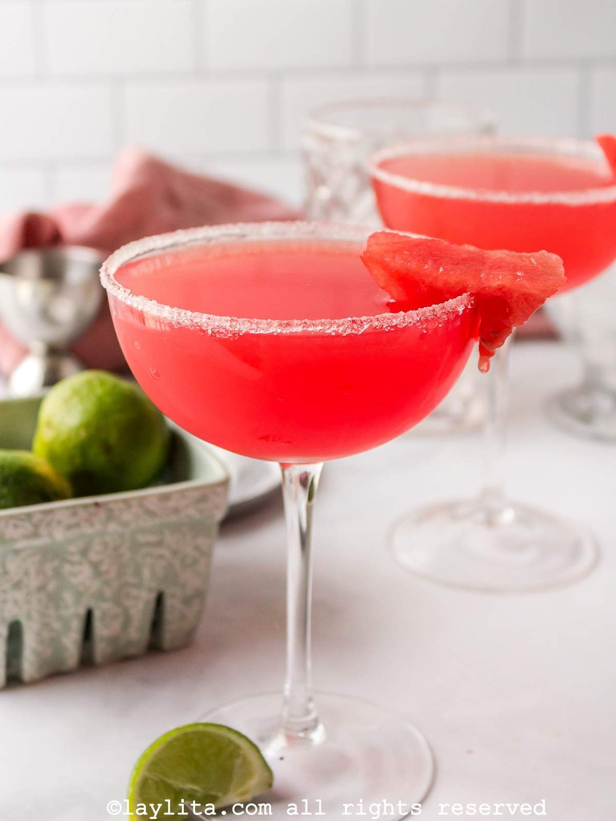 Two watermelon margaritas in coupe glasses, both garnished with sugar and watermelon, with a lime wedge and more limes on the side