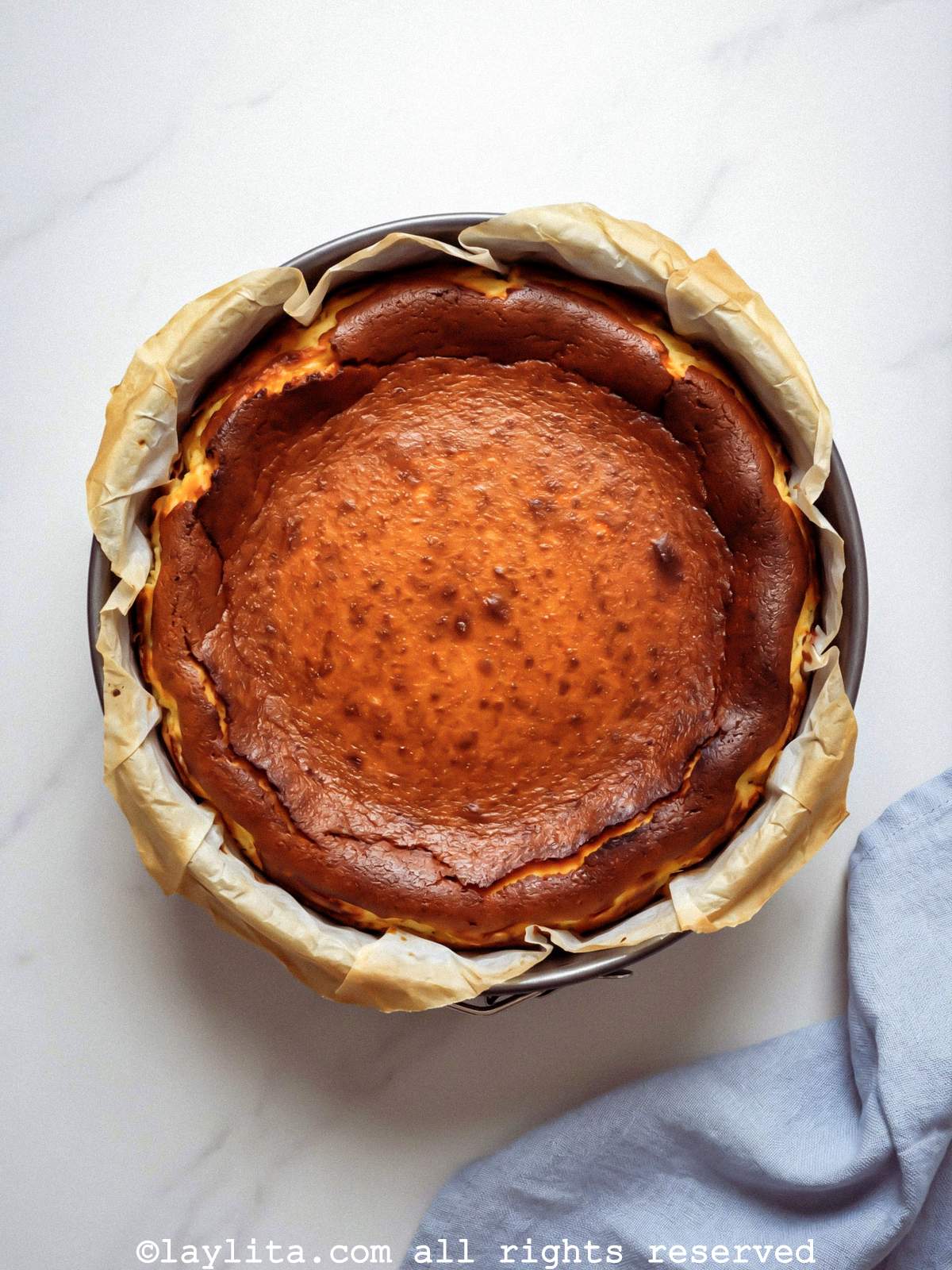 Finished baked basque cheesecake in a cake pan on a countertip next to a light blue kitchen towel