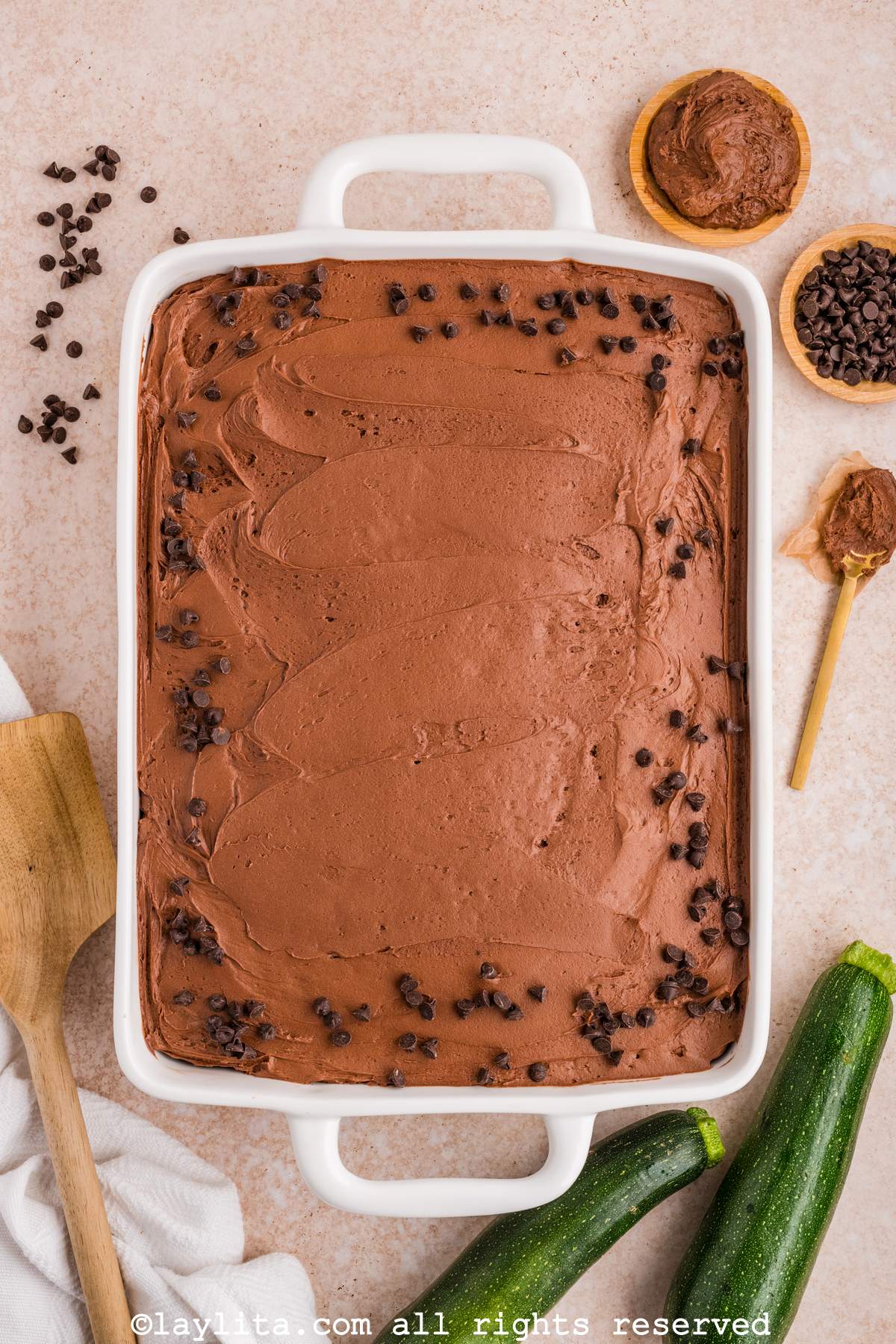 Overhead view of a white baking mold full of chocolate zucchini cake decorated with frosting and chocolate chips with a couple of zucchinis and more chocolate chips on the side