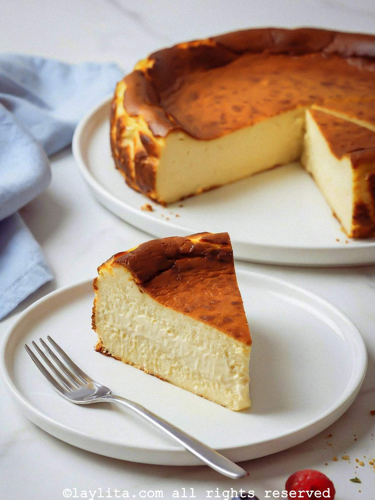 A slice of basque cheesecake on white plate with a fork, with the rest of the cheesecake in the background