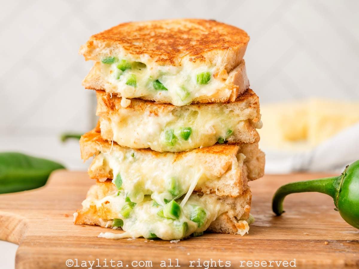Four slices of grilled cheese with jalapeños stacked on top of each other on a wooden board.