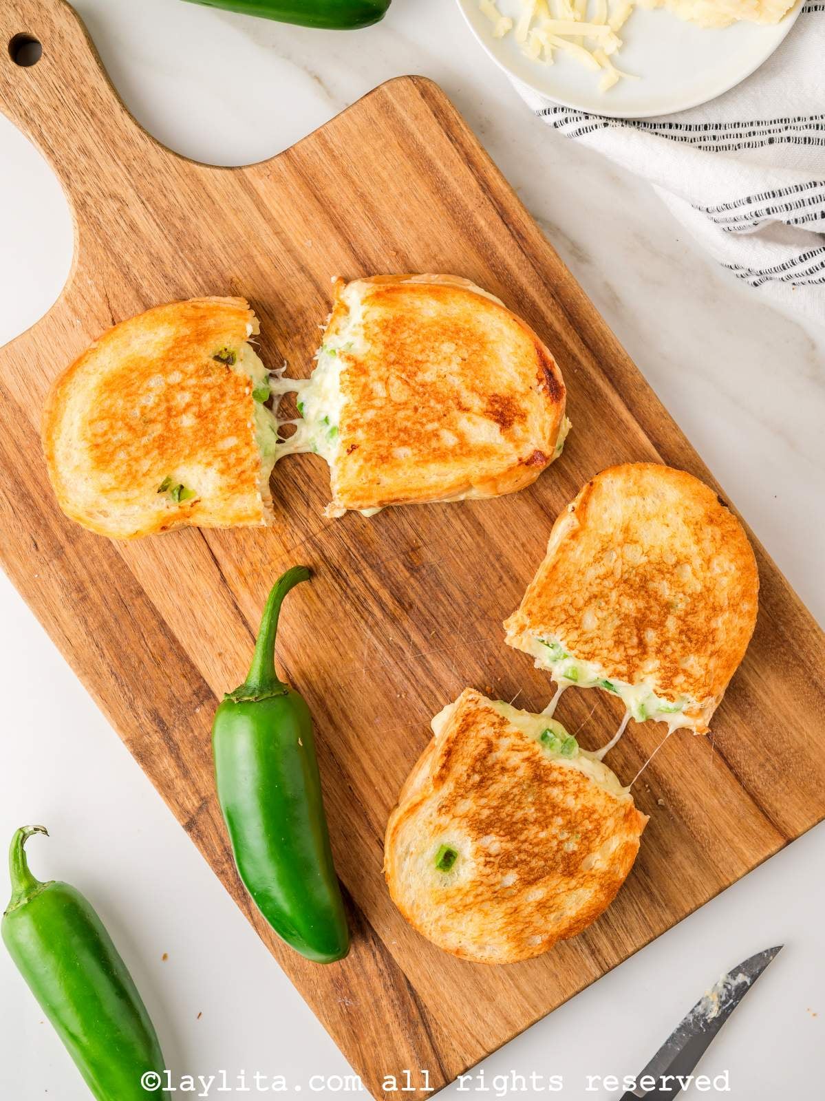 Overhead view of a two grilled cheese sandwiches cut in half on a wooden board next to a couple of jalapeño peppers