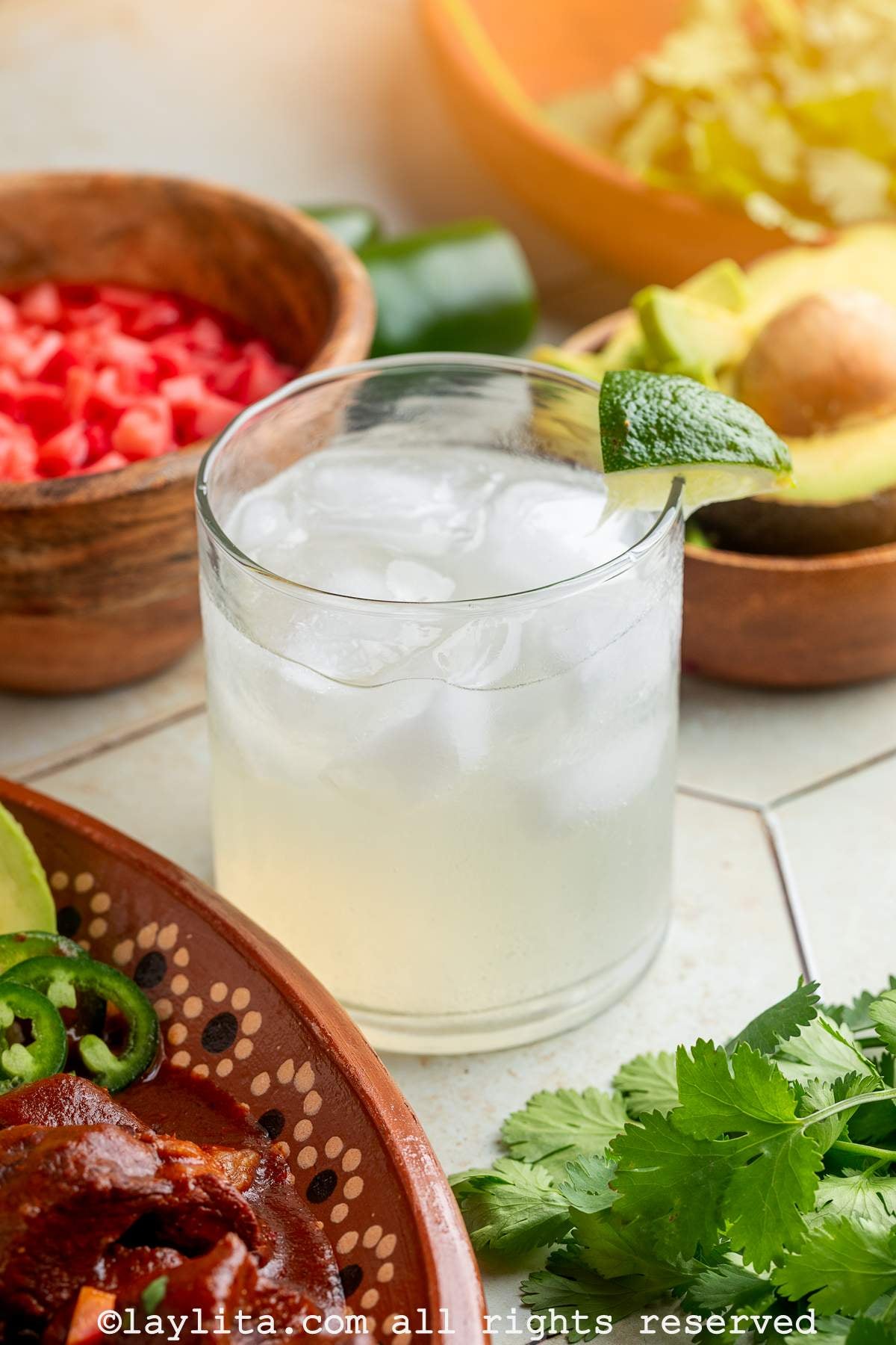 A margarita in a tumbler glass, garnished with a lime wedge, with beef stew, pickled radishes, avocado and cilantro surrounding the glass