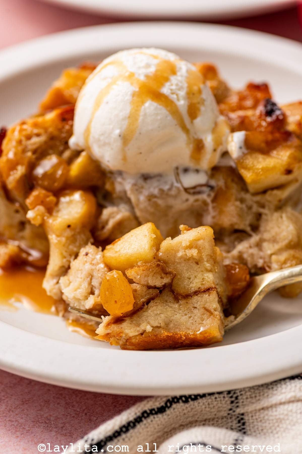 A portion of bread pudding topped with vanilla ice cream and caramel on a plate, with a forking taking a bite