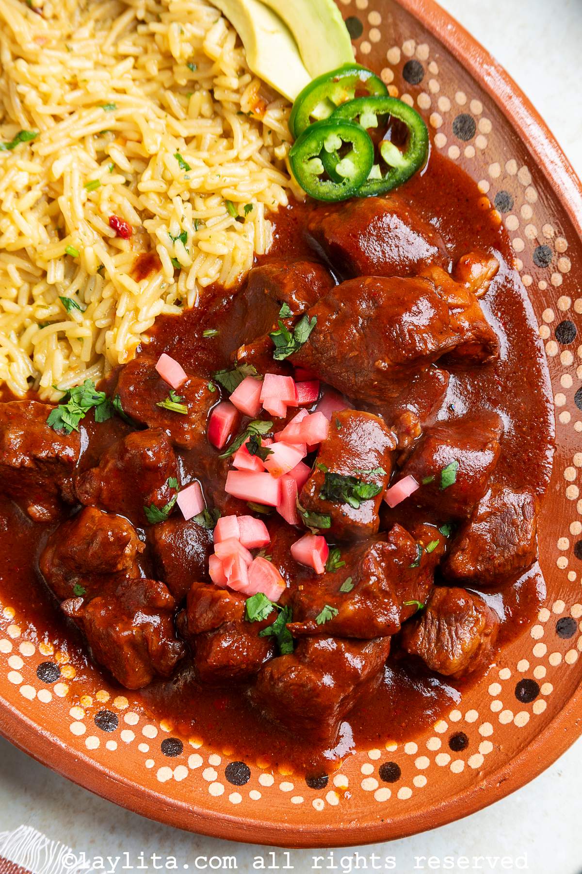 Overhead view of Beef Chile Colorado stew with rice, avocados and jalapeño slices on a terracotta plater with a floral design on the platter borders
