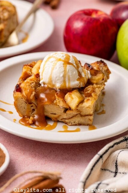 Side view of a portion of Apple Bread Pudding with Salted Caramel and a scoop of vanilla ice cream on top, with fresh apples in the background