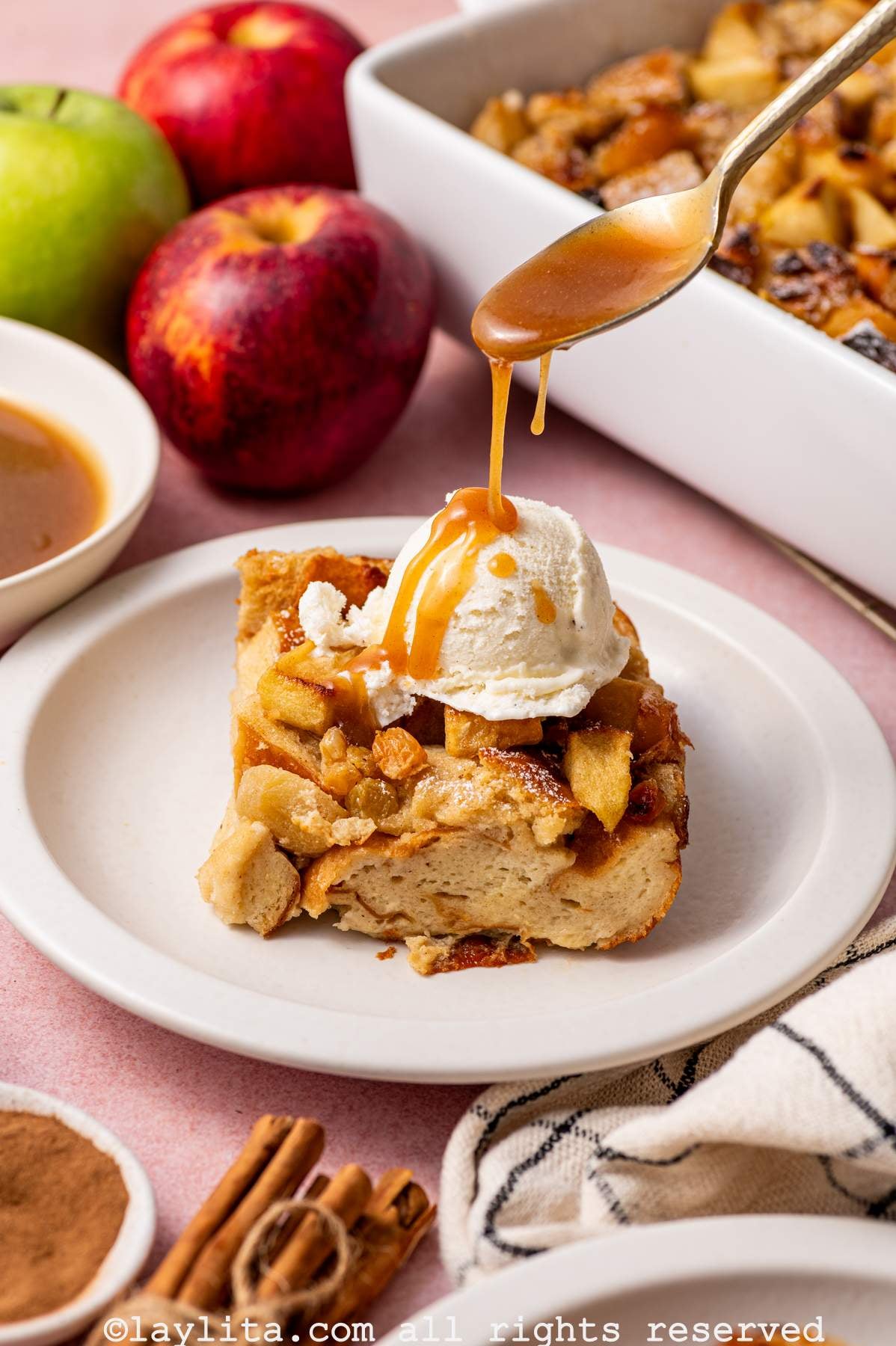Side view of a portion of apple bread pudding topped with ice cream on a plate with a spoon drizzling caramel sauce on top of it