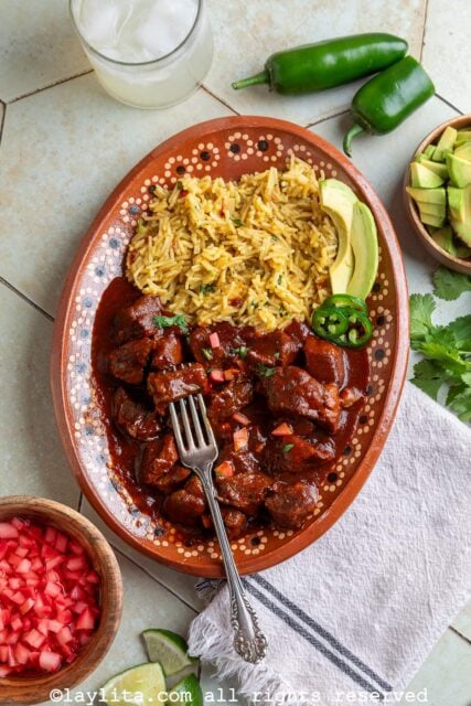 Overhead view of a platter of beef chili colorado topped with pickled radishes on a table next to a drink, whole jalapeños, more pickles, avocado, lime wedges and cilantro leaves