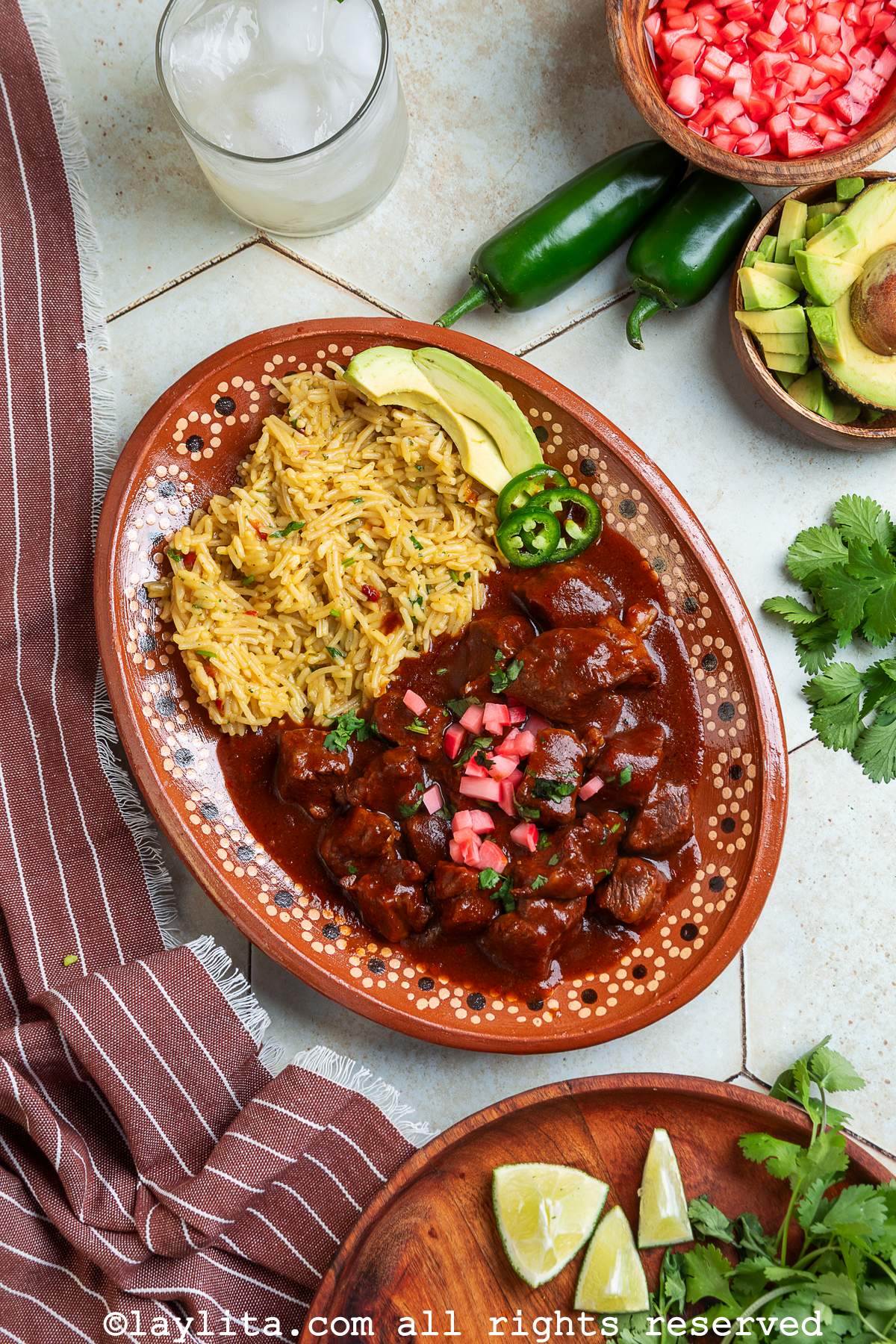 Overhead view of a platter of beef chili colorado topped with pickled radishes on a table next to a drink, whole jalapeños, more pickles, avocado, lime wedges and cilantro leaves