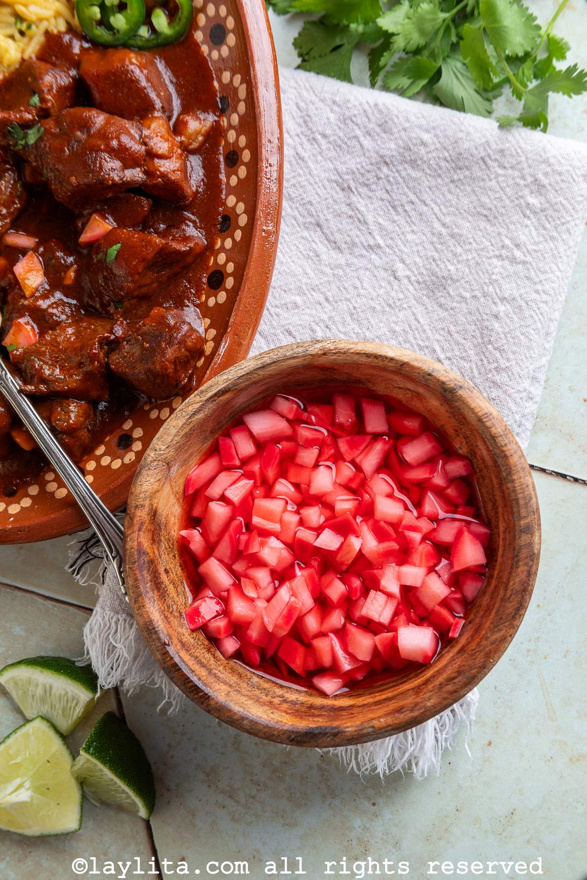 A wooden bowl with diced pickled radishes, next to a platter of beef stew and lime wedges