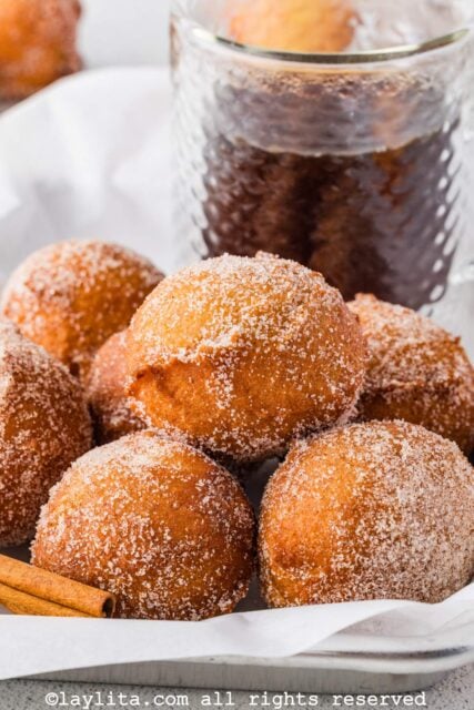 Several apple buñuelos or apple beignets in a basket with a cinnamon stick and a brown sauce in the background