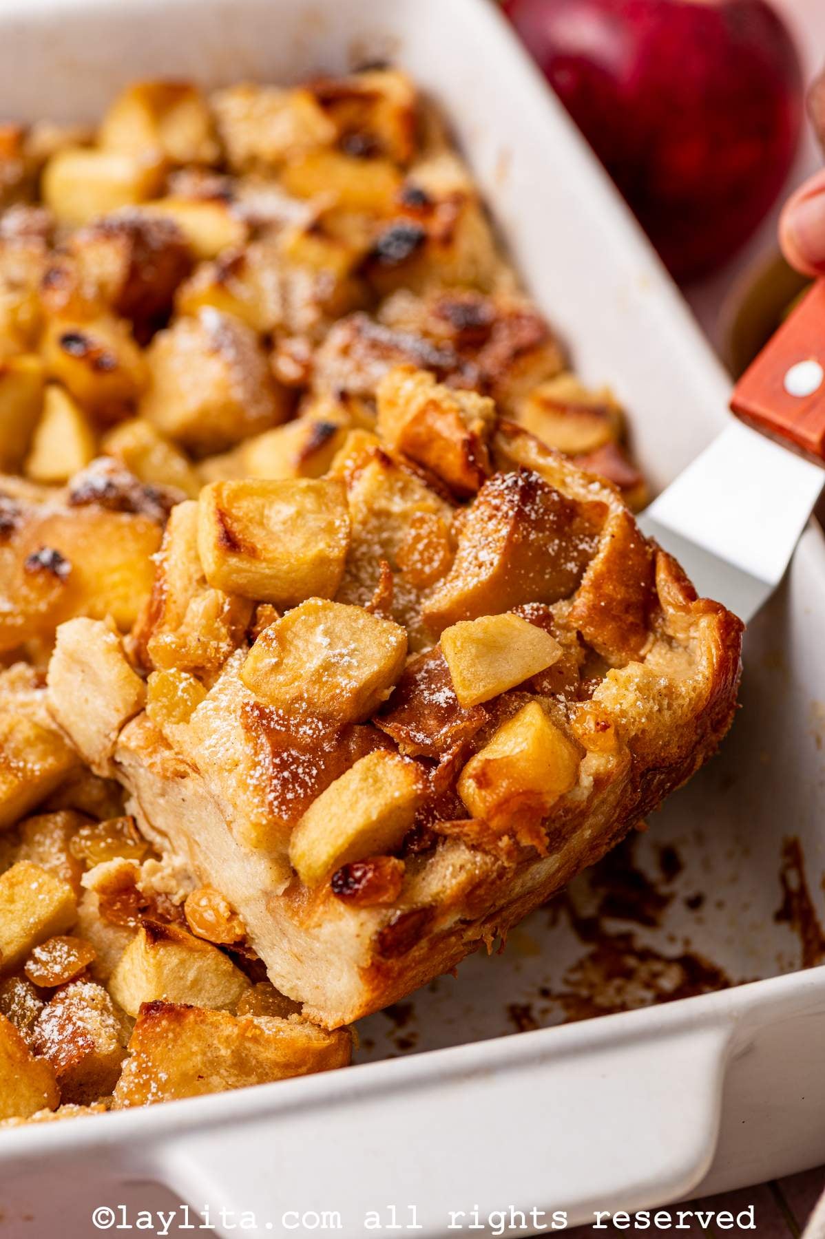 Side view of apple bread pudding in a casserole dish with a piece being lifted out with a spatula