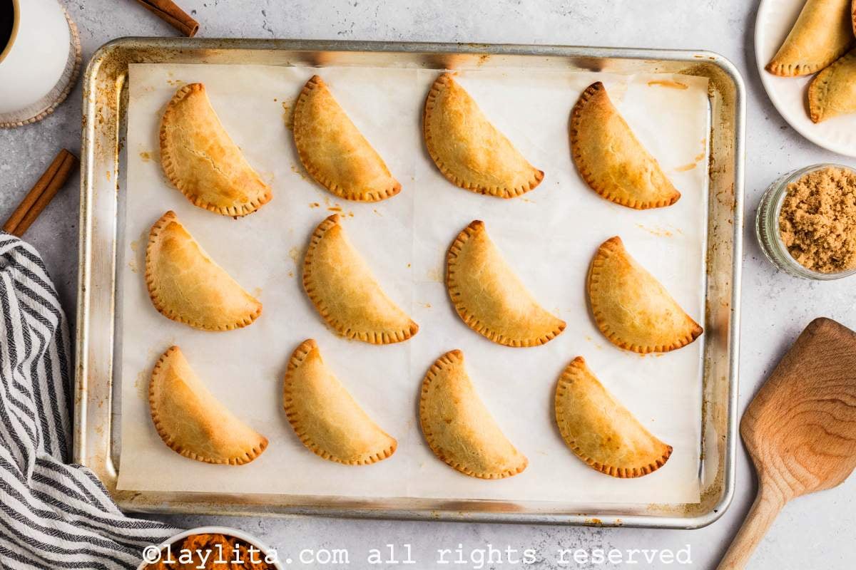 Overhead view of a tray of sweet potato empanadas, with a kitchen towel, a wooden spatula and additional empanadas on the side