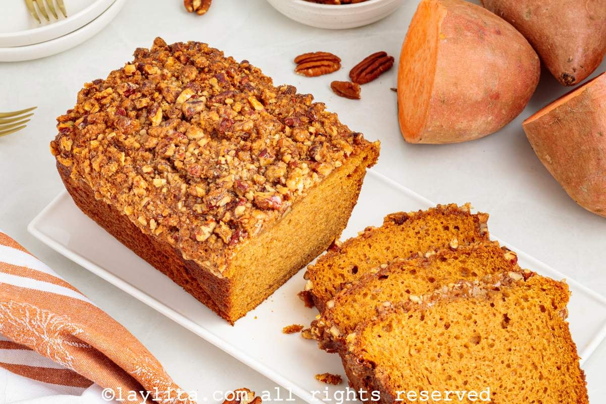 A sweet potato loaf cake topped with pecan streusel on a white plate with several pieces sliced off, in the background there are sweet potatoes and pecans