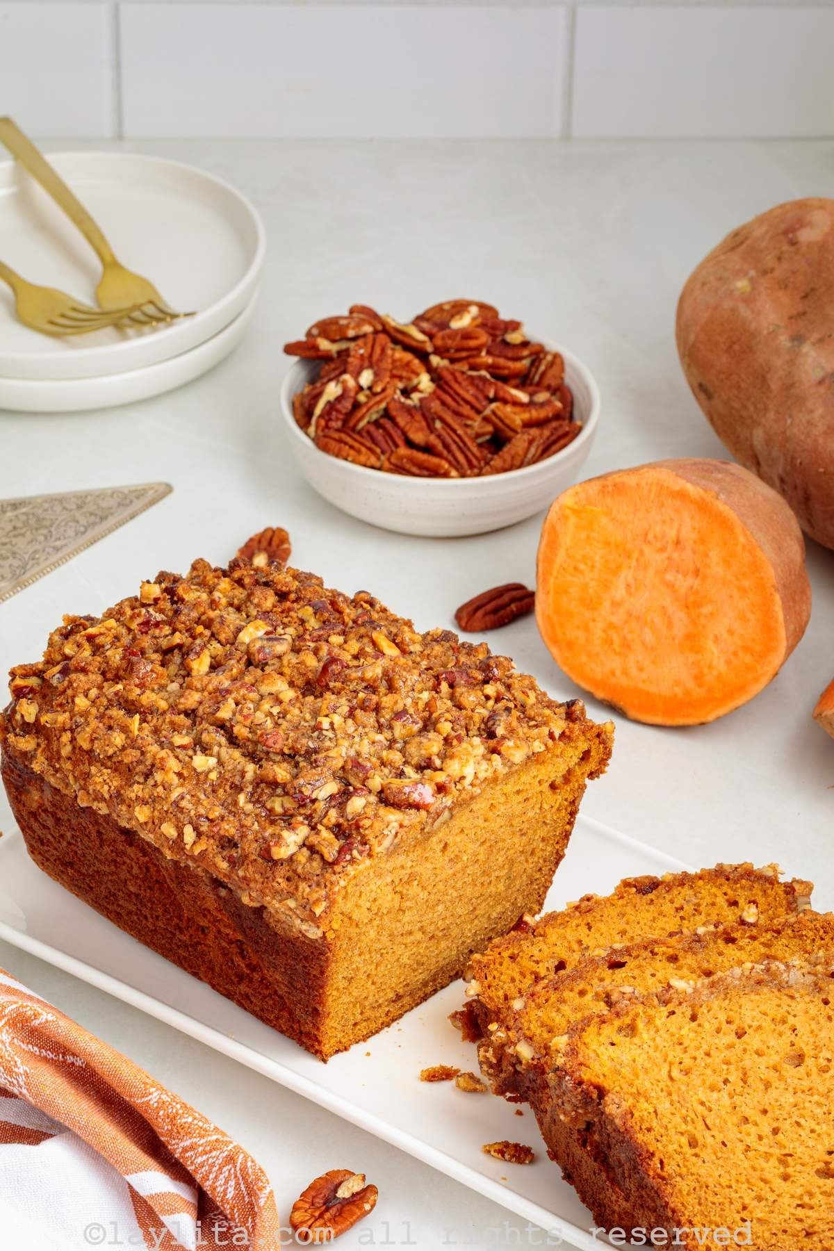A sweet potato loaf cake topped with pecan streusel on a white plate with several pieces sliced off, in the background there are sweet potatoes and pecans